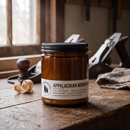 An amber glass jar candle with a black lid and a white label reading 'APPALACHIAN WORKSHOP' sits on a rustic wooden table in a workshop setting, with a hand plane and wood shavings blurred in the background.