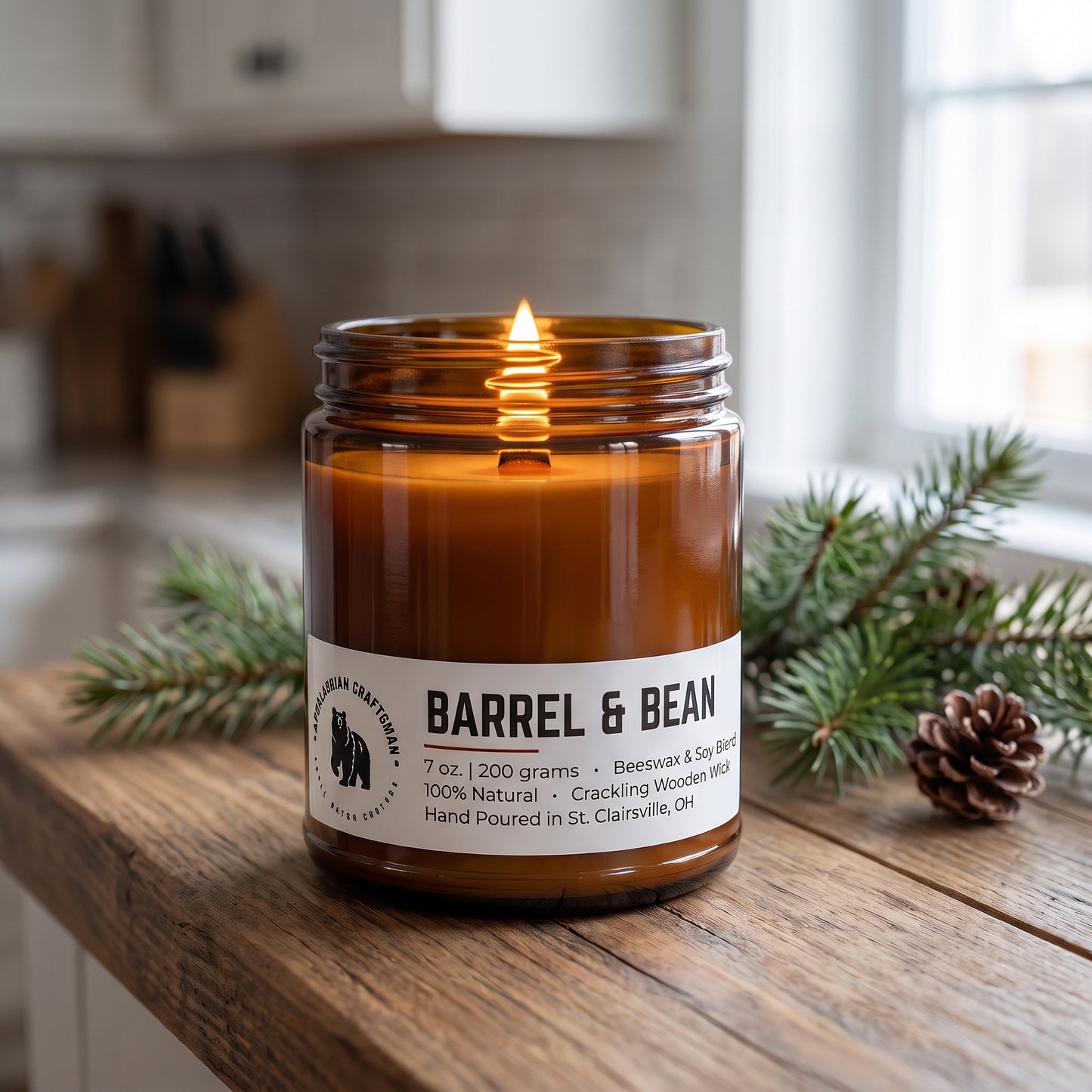 A lit amber glass jar candle sits on a rustic wooden surface with evergreen branches and a pinecone around it. The label reads 'BARREL & BEAN'.