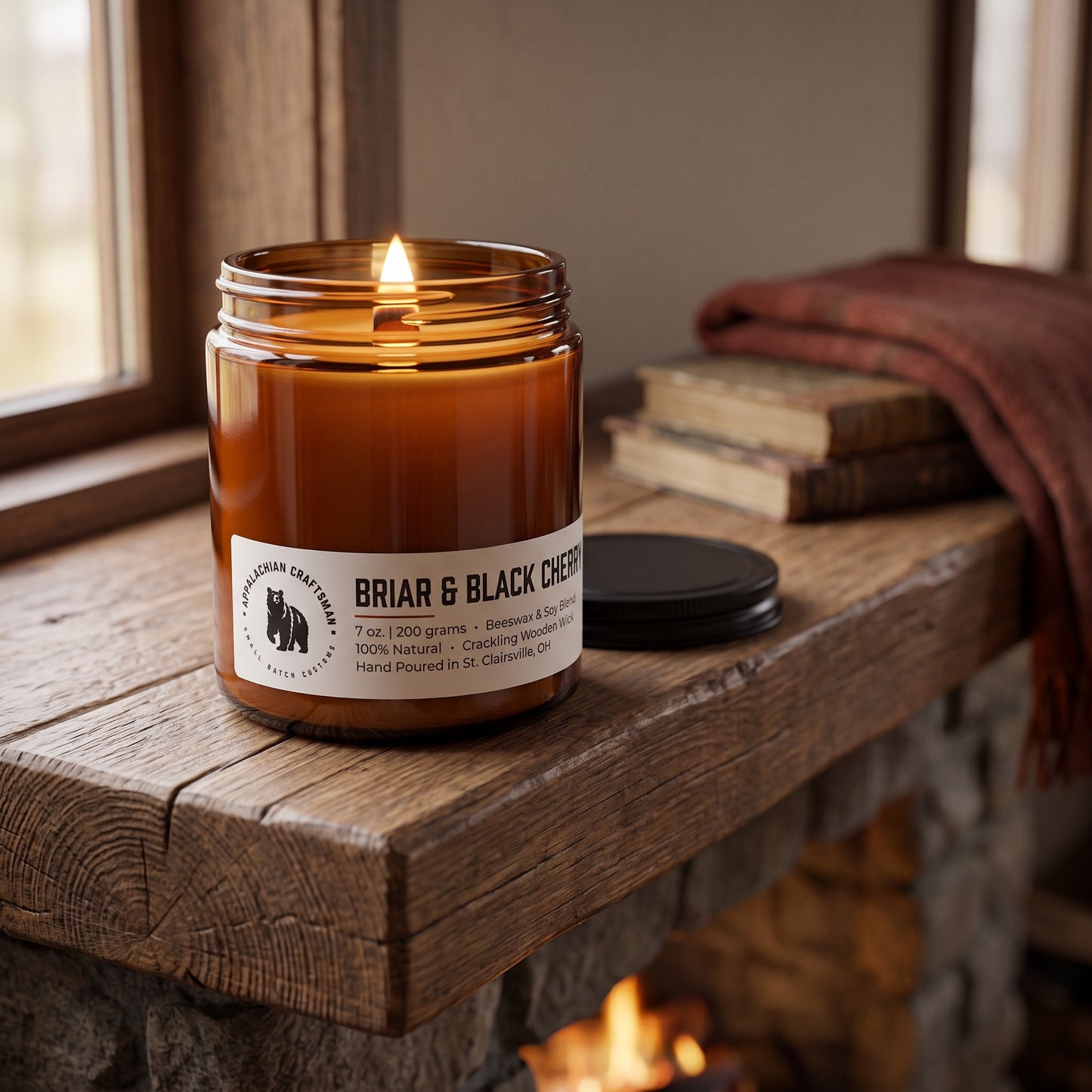 A lit amber glass jar candle labeled 'BRIAR & BLACK CHERRY' sits on a rustic wooden mantel beside a closed black lid, with stacked books and a draped red textile in the background near a fireplace.