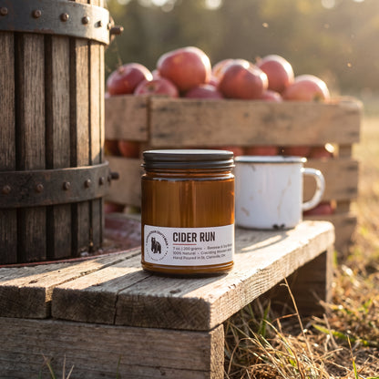 A jar candle labeled 'CIDER RUN' sits on a rustic wooden bench outdoors, with a crate of red apples and a white enamel mug blurred in the background.