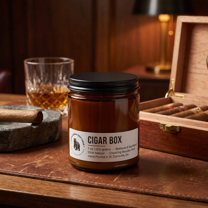 A candle jar labeled 'Cigar Box' sits on a wooden table beside an open wooden cigar box, a glass of amber liquor, and a cigar in a dimly lit bar setting.