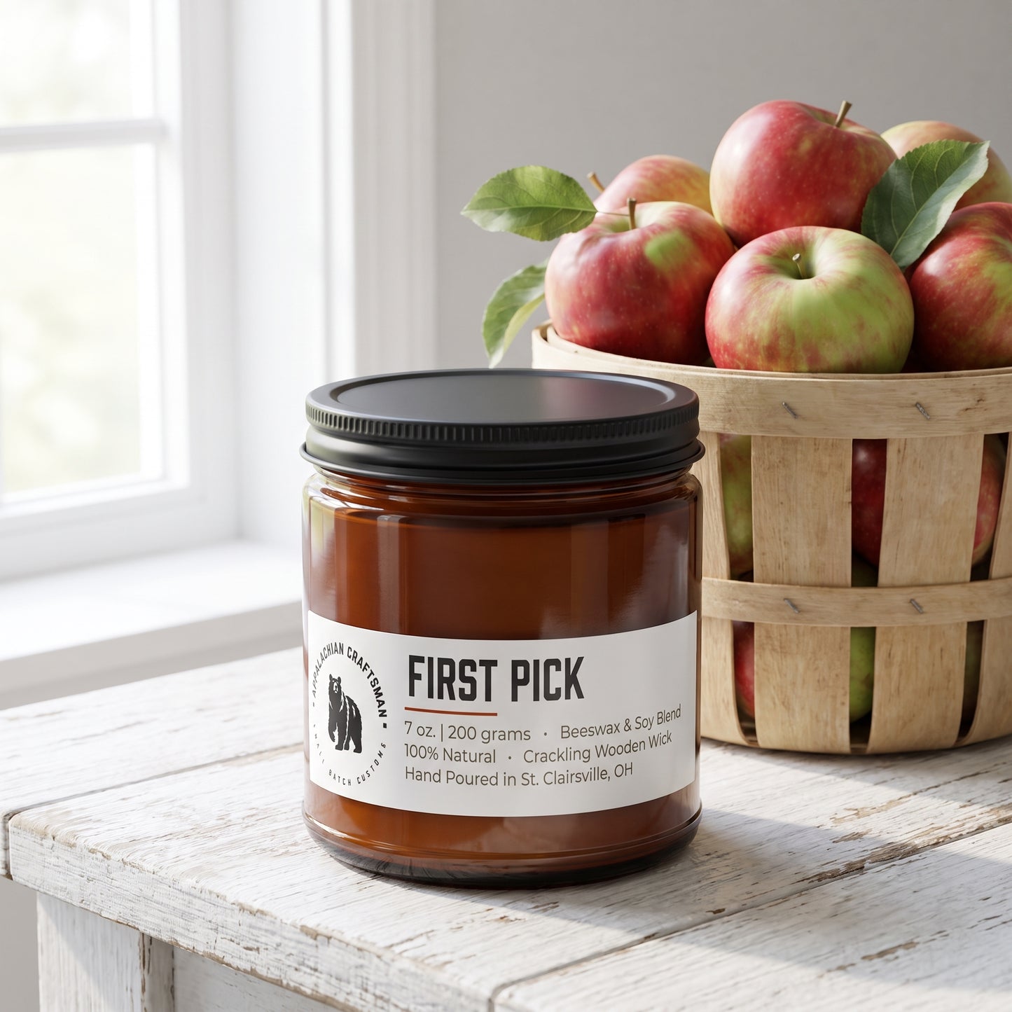 A jar candle labeled 'FIRST PICK' on a rustic white wooden table, with a basket of red and green apples in the background.