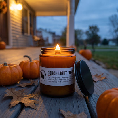 A lit amber glass jar candle labeled 'PORCH LIGHT PUMPKIN' sits on a wooden porch table with pumpkins and autumn leaves around it, with a blurred house and yard in the background.