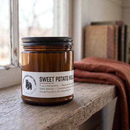 An amber glass jar candle with a black metal lid and a white label reading 'SWEET POTATO HOLLOW' sitting on a rustic wooden windowsill with a blurred background of a window, books, and a rust-colored blanket.